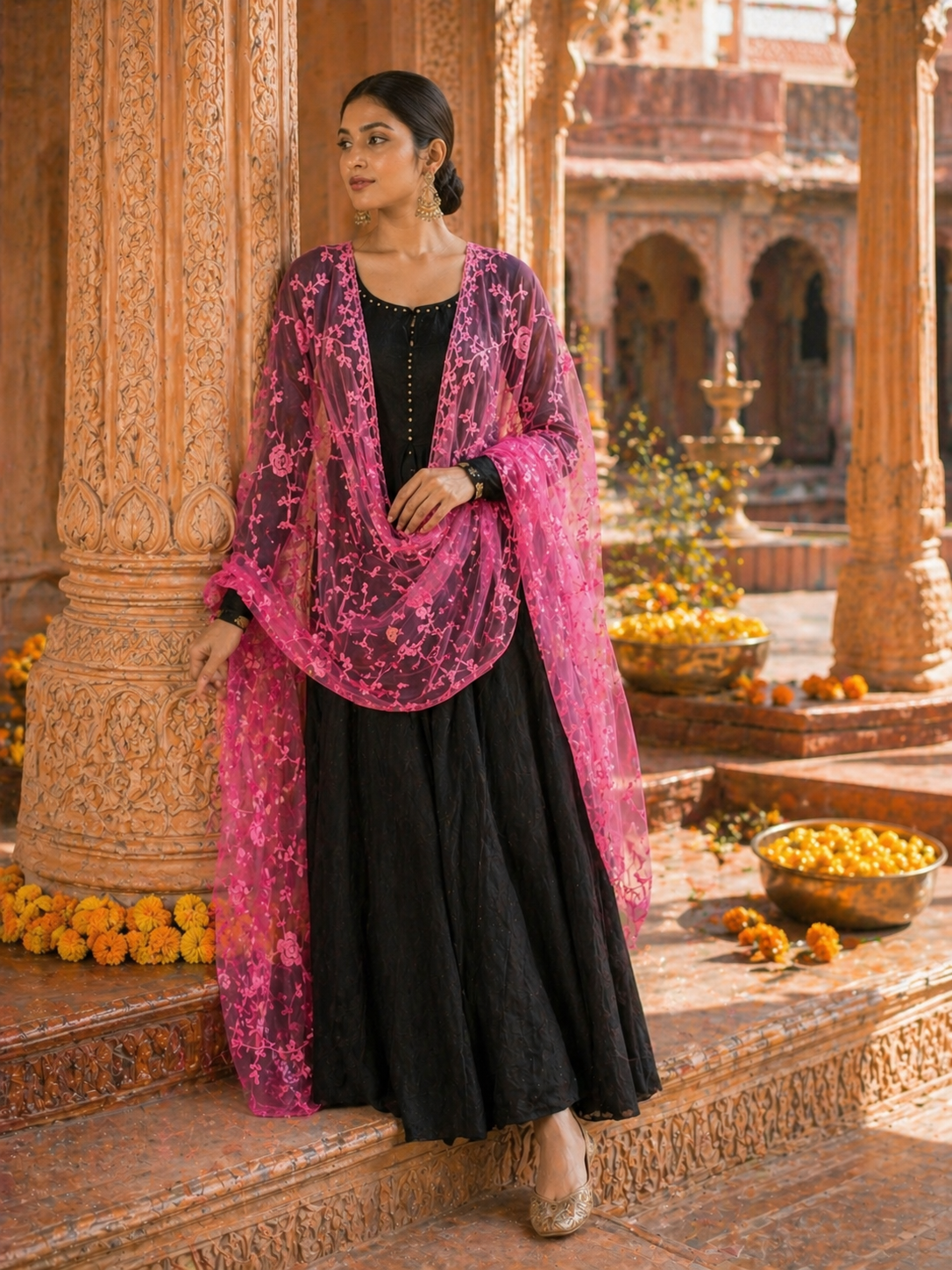 Woman in a black outfit with a pink dupatta standing in an ornate outdoor setting.