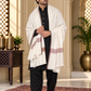 A man wearing a black traditional outfit and sandals, draped in a white shawl with red and blue geometric patterns, standing in an ornate indoor setting with a patterned rug.