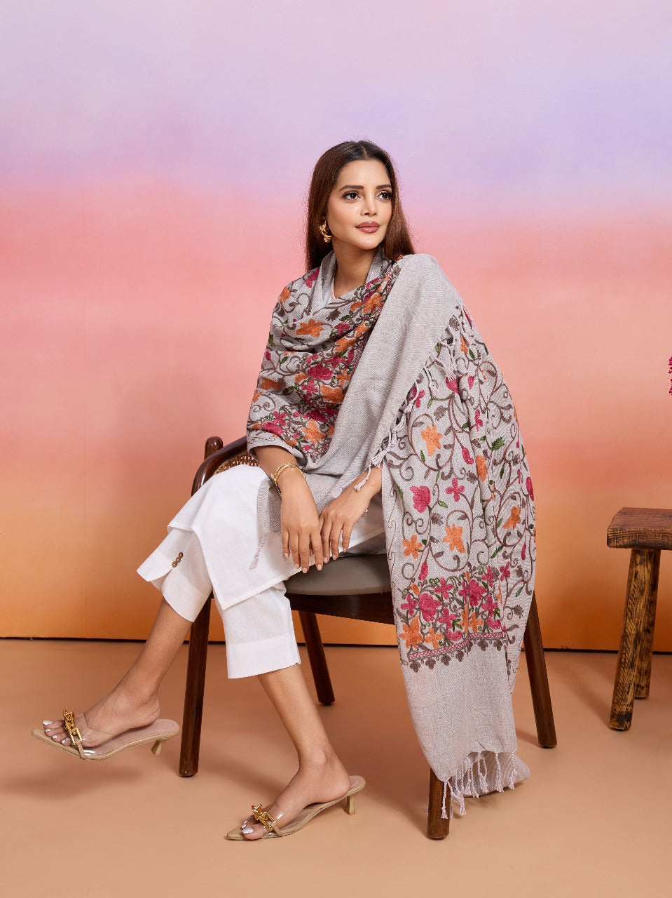 Woman sitting on a chair with a floral shawl draped over her, against a pink and orange gradient background.