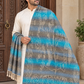 A man wearing a cream kurta stands in front of a carved wooden door, draped in a large woven shawl with horizontal stripes of turquoise blue, gray, and beige floral patterns and cream fringe.