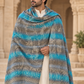 A man wearing a cream kurta and draped over his shoulders is a large patterned blue and gray shawl with fringe, standing in front of a stone archway.
