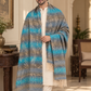 A man wearing a white kurta and trousers is draped in a large woven shawl with horizontal stripes of blue, gray, and beige, featuring fringe details, set in an elegant indoor room.