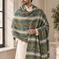 A man wearing a cream kurta and draped over his shoulders is a large, textured striped shawl or wrap in shades of green, olive, and beige with fringed edges, set in a bright, modern indoor environment with marble walls and a window.