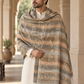 A man wearing a cream kurta and a large striped shawl with beige, brown, and gray tones and subtle floral motifs, standing in an arched corridor.