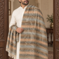 A man wearing a white kurta stands in an ornate interior doorway, draped with a large patterned shawl in beige, brown, gray, and muted blue tones with fringe tassels.
