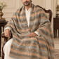 A man seated in an ornate armchair wearing a white kurta and draped over his shoulders is a large patterned shawl or throw blanket in beige, brown, gray, and muted blue tones with fringe at the ends, in an elegant indoor setting.