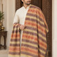 A man wearing a cream kurta and draped a striped multicolor shawl with fringe, standing in front of an ornate wooden door.