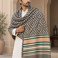 A man wearing a white kurta draped with a large woven shawl in a beige courtyard. The shawl features a gray and beige geometric pattern with a decorative border in green, peach, and beige, finished with cream fringe.