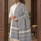 A man wearing a cream kurta and white trousers stands in an ornate indoor setting, draped in a large gray and cream patterned shawl with blue stripes and fringe.