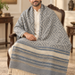 A man seated in an ornate wooden armchair wearing a cream kurta and draped over his shoulders is a large woven shawl or throw blanket with a blue and cream geometric pattern and fringe trim, in a traditional Indian living room setting.