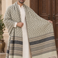 A man wearing a cream kurta and trousers models a large woven shawl with a beige and gray geometric pattern and striped borders, standing in front of a carved wooden door.