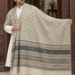 A man wearing a white kurta and trousers models a large woven shawl draped over his shoulders. The shawl features a beige and gray chevron pattern on the upper section, a wide dark gray patterned band across the middle, and cream-colored fringe along the bottom.