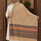 A man wearing a cream kurta stands in a doorway holding up a large patterned shawl. The shawl is beige with a geometric diamond pattern and a wide border featuring stripes of rust orange, brown, and dark blue, finished with white fringe at the bottom.