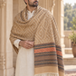 A man wearing a cream kurta and trousers, draped in a large beige patterned shawl with orange and dark brown geometric borders and tassels, standing in front of an ornate stone archway.