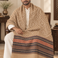 A man seated in a wooden chair wearing a beige patterned shawl over a white kurta and trousers, with the shawl draped over his lap in a warm indoor setting.
