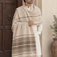 A man wearing a cream-colored kurta and trousers models a large beige and brown patterned woolen shawl with fringe, standing in front of a rustic wooden door and a potted plant.