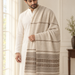 A man wearing a white kurta and white trousers, draped with a large cream and brown striped woolen shawl or throw blanket with fringe, standing in a bright indoor room.