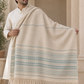 A man wearing a cream kurta holds up a large cream shawl or wrap with light blue and beige horizontal stripes and fringe along the bottom, posed in front of a beige stone archway.