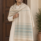 A man wearing a cream-colored kurta and matching trousers, draped with a large cream shawl featuring light blue stripes and fringed edges, standing in front of a wooden door.