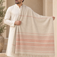 A man wearing a cream kurta stands in front of a beige arched doorway, holding up a large cream shawl with thin coral-pink horizontal stripes and fringe along the bottom edge.