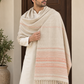 A man wearing a cream kurta and matching trousers is draped in a large woven shawl with peach and cream horizontal stripes and fringed edges, standing in front of a dark wooden door.