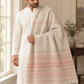 A man wearing a white kurta and matching trousers, draped with a cream-colored shawl featuring soft pink stripes and fringed edges, standing in a bright, neutral-toned indoor setting.