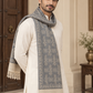 A man wearing a cream-colored kurta and a gray patterned shawl scarf with beige fringe, standing in an elegant indoor setting.