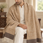 A man seated indoors wearing a beige patterned shawl with cream fringe and dark brown border panels over a white kurta and trousers.