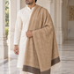 A man wearing a white kurta and trousers models a large beige and brown patterned shawl with white fringe, standing in a grand marble corridor with columns.