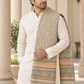 A man wearing a cream kurta and a beige patterned shawl with orange and brown woven stripes, standing in front of a stone columned building.