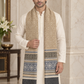 A man wearing a cream kurta and white trousers models a long woven scarf with beige, blue, and gray geometric patterns and fringe, set against an elegant marble interior background.