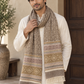 A man wearing a cream kurta and a patterned woolen scarf in beige, brown, and muted pink, standing in front of a beige wall and dark wooden door.