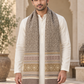 A man wearing a long cream and brown patterned knit scarf with fringe, draped over a white kurta, standing in front of a beige architectural background with arches.