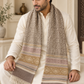 A man wearing a cream-colored kurta sits on a beige sofa, draped with a thick, patterned woolen scarf in neutral shades of beige, brown, and muted pink. The scarf features geometric and floral motifs with fringed ends.