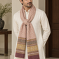 A man wearing a cream-colored kurta and a dusty pink patterned scarf with multicolor geometric borders and tassels, standing in an indoor setting.